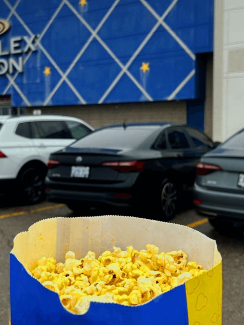 Four Volkswagens are parked next to eachother in the parking lot of a Cineplex movie theatre. In the foreground is a blue bag of movie-theatre popcorn.
