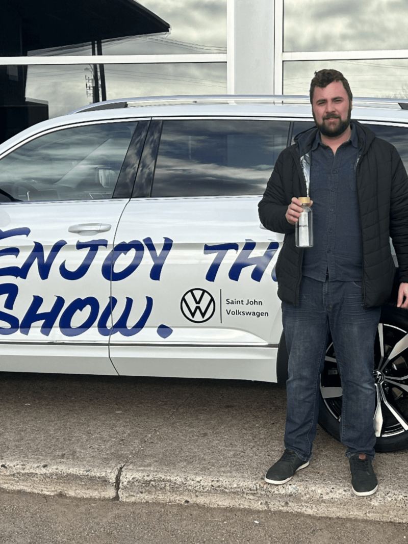 Two men are standing in front of a parked Volkswagen SUV with the trunk open. One of them has the Volkswagen logo on his jacket. On the side of the car, there is a decal that reads, “Enjoy the show.” 