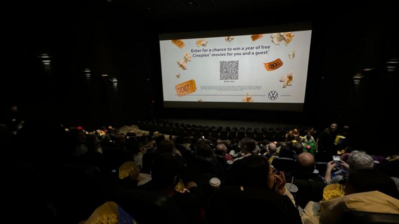 A movie theatre auditorium full of people. On the screen, there is a barcode surrounded by popcorn kernels and yellow movie ticket stubs. Above it reads, “Enter for a chance to win a year of free Cineplex® movies for you and a guest.” The Volkswagen logo sits in the bottom right corner of the screen. 