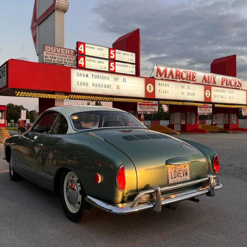 A VW Karmann Ghia coupe with a ‘Love VW’ license plate is parked outside a drive-in movie theatre. 