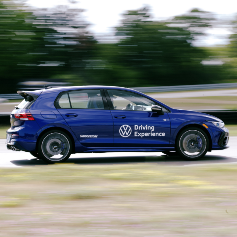 A blue Golf R driving on a racetrack with a VW Driving Experience decal on the side. The background is motion-blurred. 