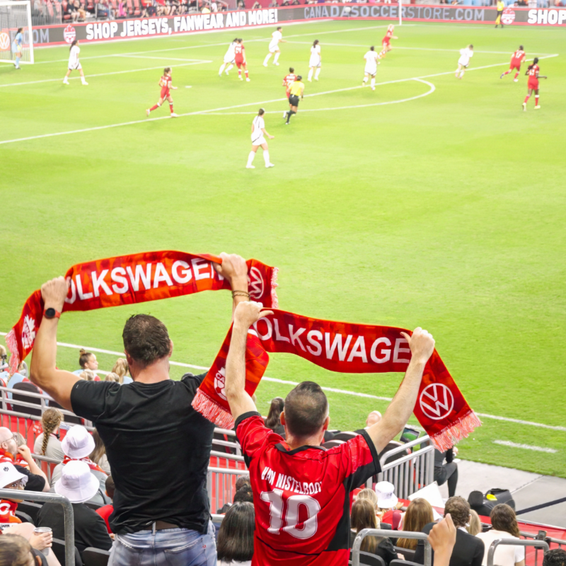 Shot from the stadium stands during the Canada Women’s Soccer Pride Match, with two fans holding up VW × Team Canada scarves. 