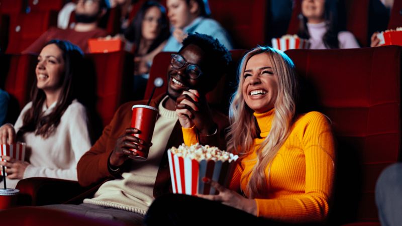 Deux personnes sont assises dans une salle de cinéma et sourient devant l’écran. L’un mange du pop-corn dans un grand seau jaune.