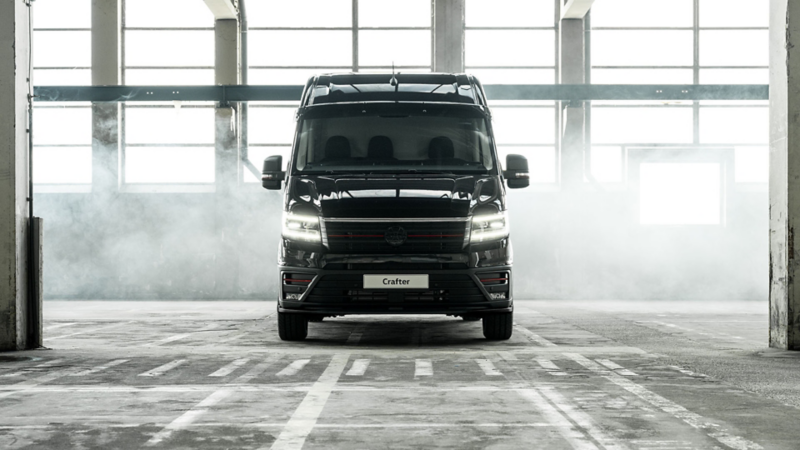 Vooraanzicht van een zwarte Volkswagen Crafter met brandende koplampen in een lichte, industriële hal met rook.