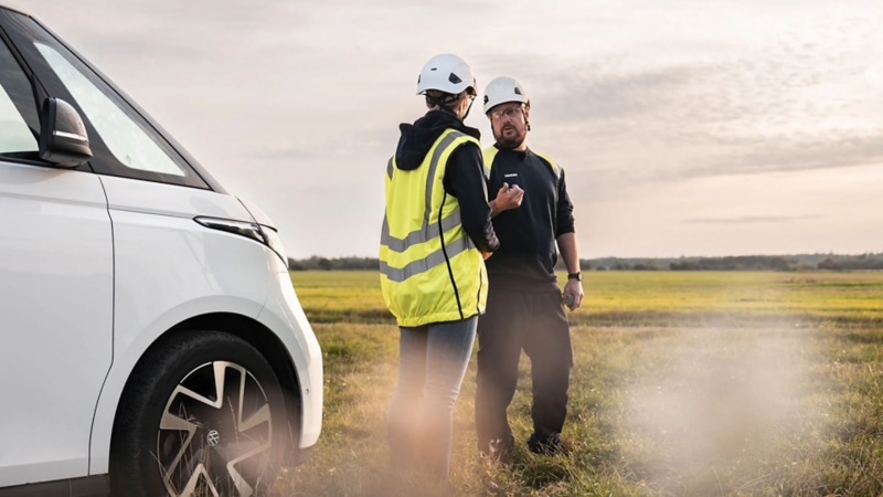 Twee professionals in veiligheidskleding praten naast een witte Volkswagen ID. Buzz in een open veld.