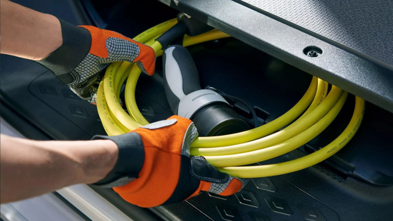 Iemand met werkhandschoenen plaatst een opgerolde gele laadkabel in de opbergruimte van een elektrische auto.