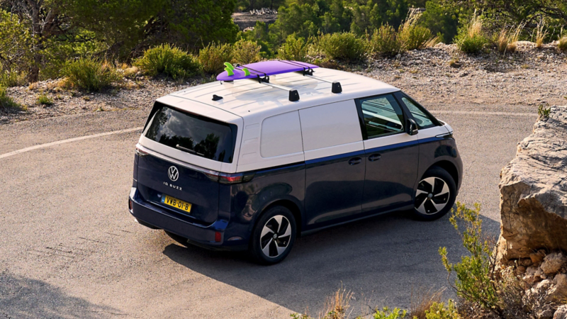 Achteraanzicht van een tweekleurige Volkswagen ID. Buzz Cargo met een surfplank op dakdragers, rijdend op een bergweg.