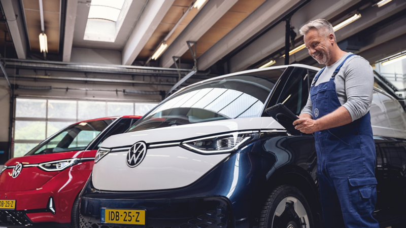 Een monteur in blauwe overal voert met een tablet een accucheck uit bij een zwart-witte Volkswagen ID. Buzz Cargo in een werkplaats.
