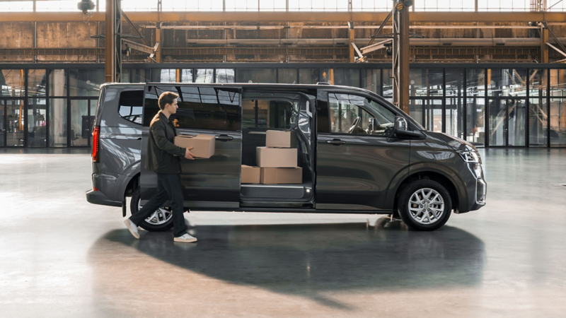 Een man laadt kartonnen dozen in de open schuifdeur van een grijze Volkswagen Caravelle eHybrid toCargo in een loods.