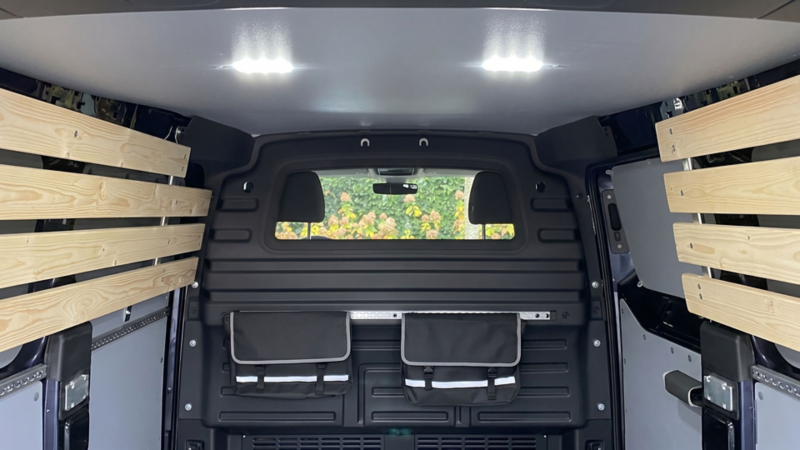 Afgewerkte laadruimte met grijze hemelbekleding, ledverlichting, een scheidingswand met ruit en houten latbekleding.