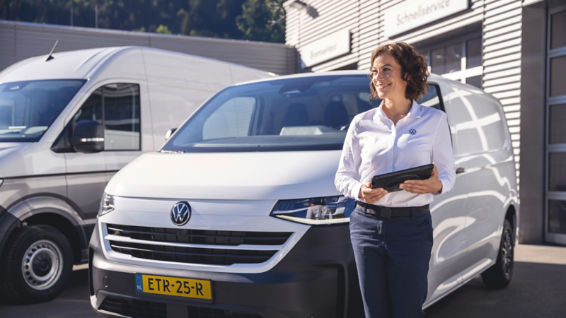 Glimlachende Volkswagen-medewerkster met een tablet in haar handen voor twee witte bedrijfswagens bij een dealer.