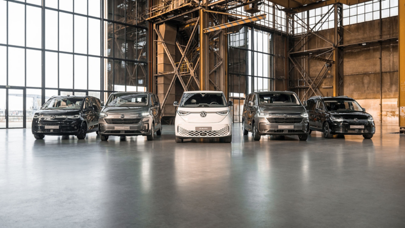 Vijf verschillende modellen Volkswagen bedrijfswagens, waaronder de witte ID. Buzz in het midden, geparkeerd in een ruime industriële hal.