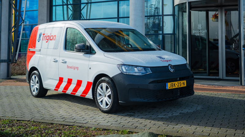 Witte Volkswagen Caddy Kombi eHybrid toCargo met Trigion Beveiliging logo en rood-witte striping geparkeerd voor een glazen entree.