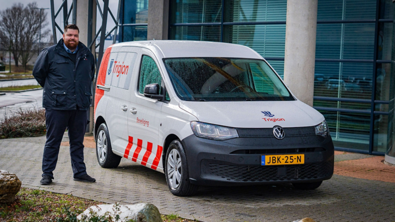 Een medewerker van Trigion Beveiliging poserend naast een witte Volkswagen Caddy Kombi eHybrid toCargo met bedrijfslogo.