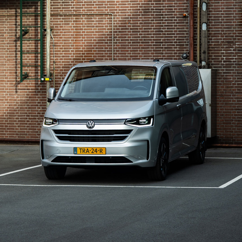 Zilverkleurige Volkswagen Transporter geparkeerd in een wit omlijnd parkeervak voor een bakstenen muur.
