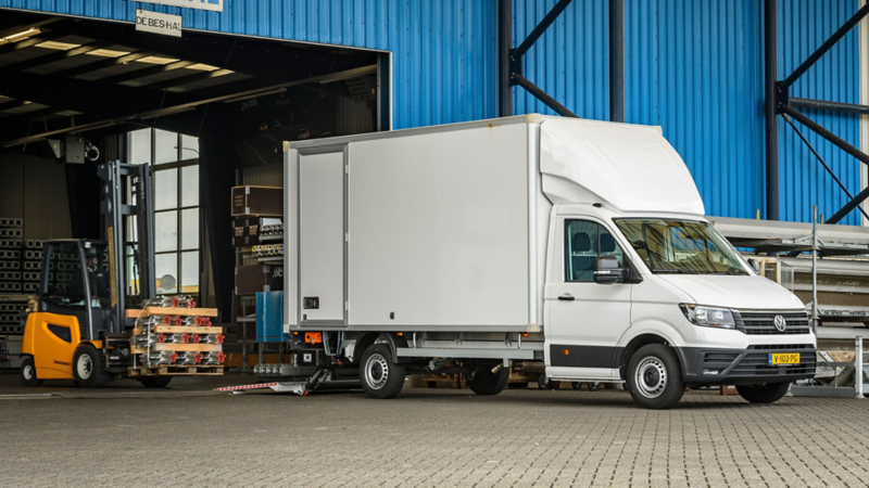 Een witte Volkswagen Crafter bakwagen staat geparkeerd voor een magazijn, met een werkende vorkheftruck op de achtergrond.