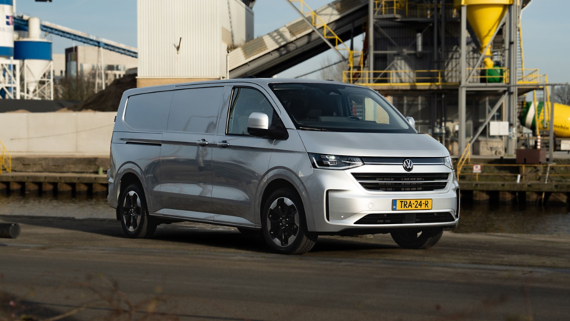 Zilveren Volkswagen Transporter geparkeerd op een kade met een industriële installatie op de achtergrond.