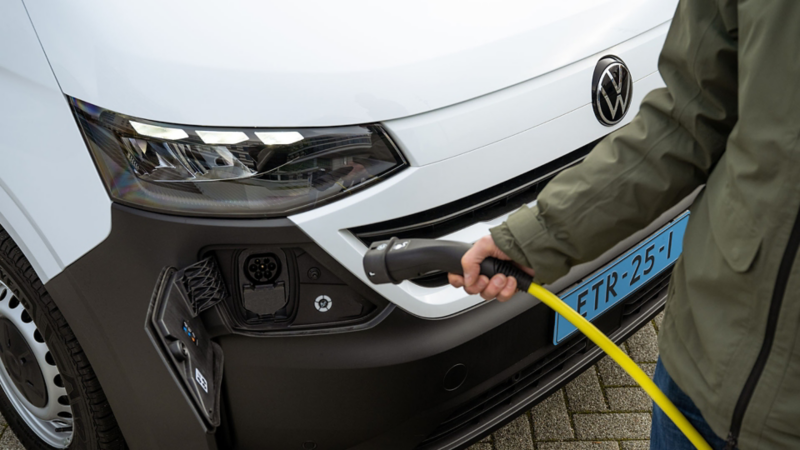 Een persoon steekt een gele laadkabel in de aansluiting van een witte elektrische Volkswagen bedrijfswagen.
