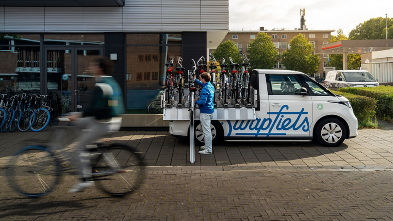 De ID. Buzz Cargo van de zijkant. Een 'Swapper' in herkenbare Swapfiets-outfit is bezig een blauwband-fiets in het rek te schuiven.
