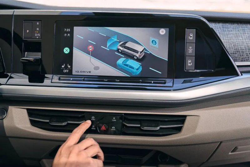 Een vinger drukt op de Assist-knop op het dashboard. Het scherm erboven toont de weergave van het IQ.DRIVE rijassistentiesysteem.