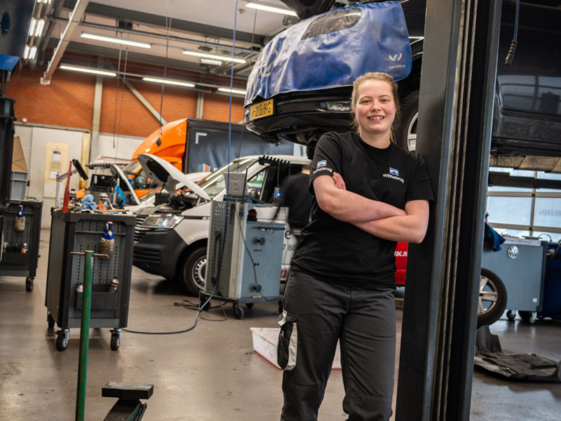 Een vrouwelijke autotechnicus staat lachend in een garage vol Volkswagen bedrijfswagens en gereedschap.