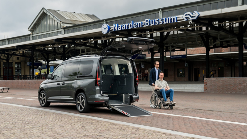 Caddy Kombi eHybrid Rolstoelauto met op de achtergrond een station en een man die een andere man in een rolstoel aan het duwen is