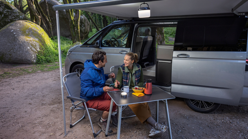 Nieuwe Volkswagen California met twee mensen die buiten zitten op klapstoelen aan een klaptafel