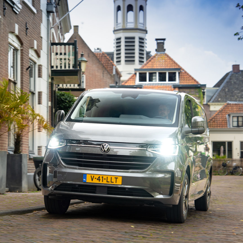 Grijze Volkswagen e-Transporter met brandende koplampen rijdt door een straat met oude panden.