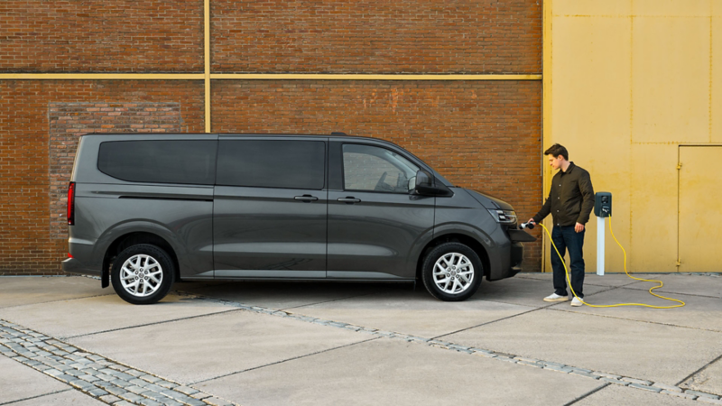 Een man sluit een grijze Volkswagen Transporter Kombi eHybrid toCargo aan op een laadpaal met een gele kabel.