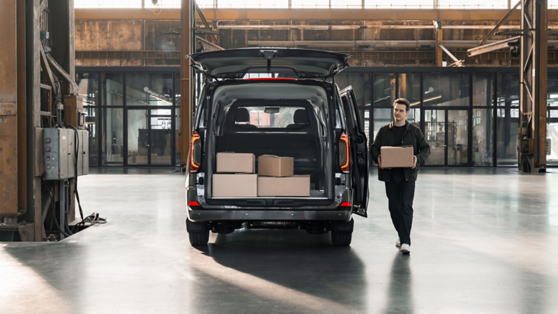 Een man loopt met een doos naar de geopende achterklep van een grijze Transporter Kombi eHybrid toCargo met een volle laadruimte.
