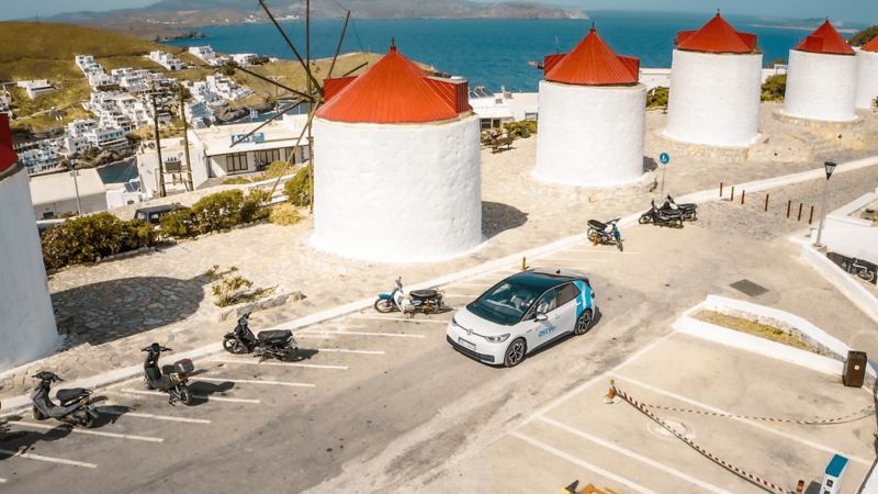 Ένα αυτοκίνητο VW-astyGO κινείται σε δρόμο μπροστά από ανεμόμυλους στην Αστυπάλαια- υπηρεσίες “astyMOVE” -Kosmocar-Volkswagen-Greece