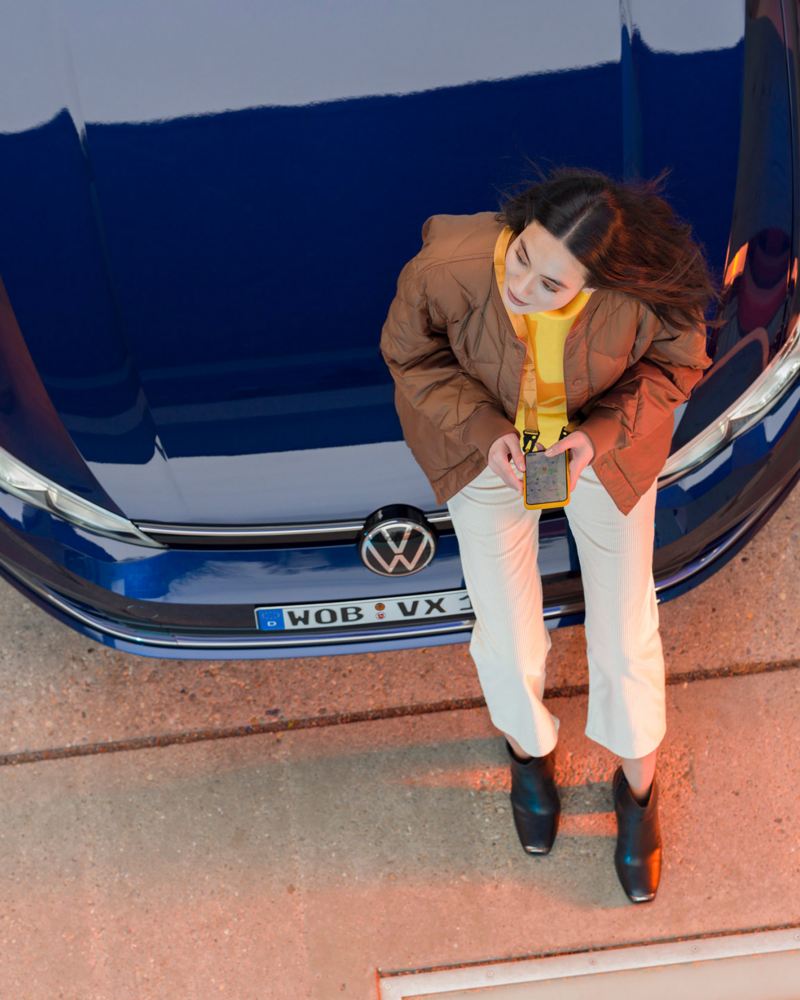Eine Frau lehnt an ihrem blauen VW mit ihrem Smartphone in der Hand – VW Apps
