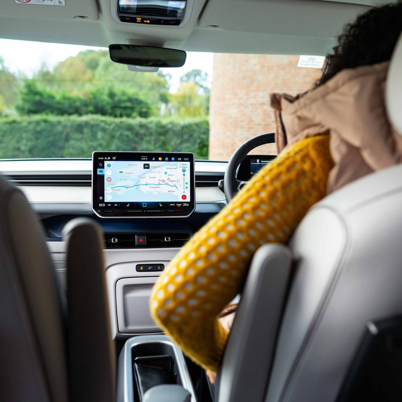 View from the back seat of a Volkswagen van showing the dashboard navigation screen and a passenger sitting in the front seat.