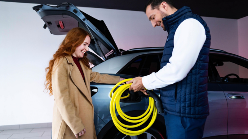 A man and woman standing next to a VW, with the man holding a charger cable