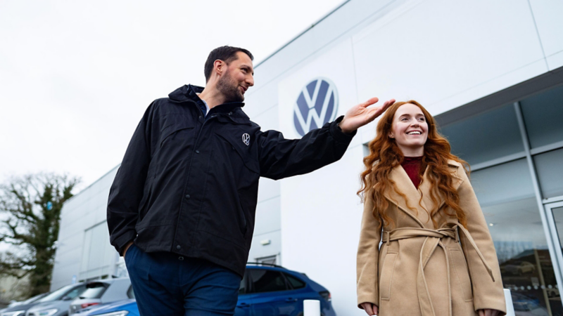 A man and woman happily walk outside a Volkswagen dealership. The man gestures animatedly, and the woman smiles warmly, conveying a friendly and positive mood.