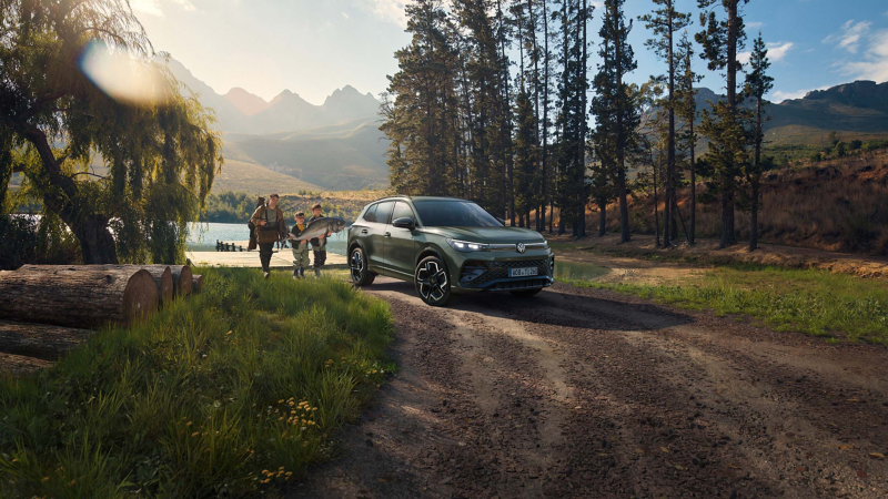 Volkswagen Tiguan SUV parkt an einen naturbelassenen Waldweg am Seeufer, umgeben von Bäumen und Bergen, während eine Gruppe von Personen mit einem Fisch in den Armen sich dem Fahrzeug nährt.