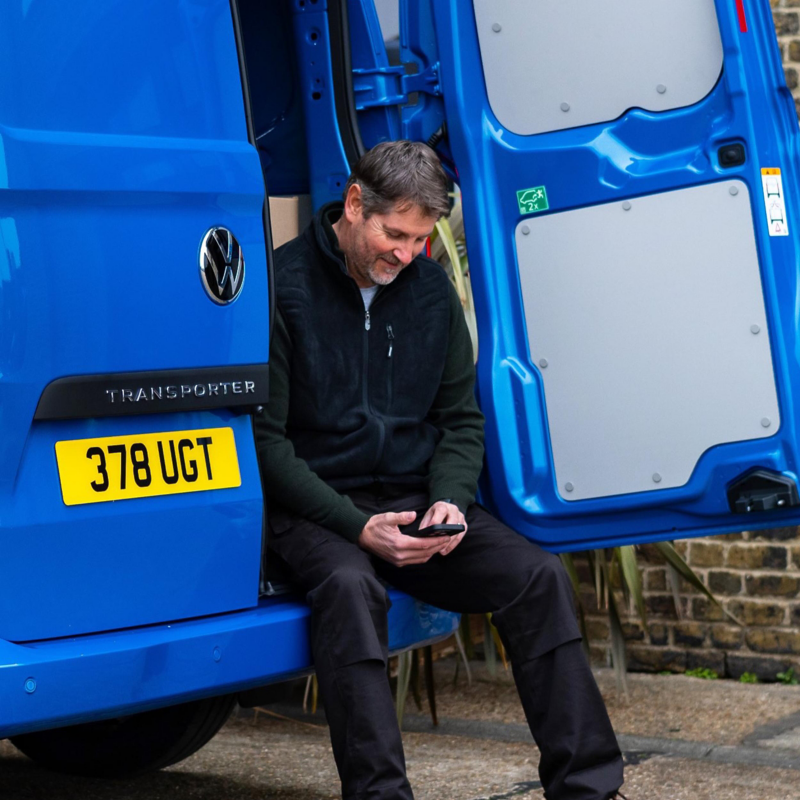 Person sitting on the rear step of a Volkswagen Transporter with the back doors open, using a smartphone.