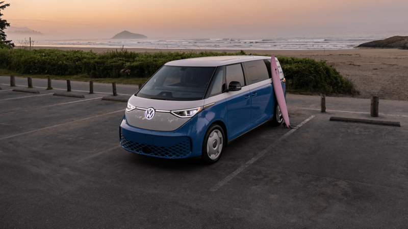 Vue trois quarts avant d'un Volkswagen ID. Buzz bleu et blanc garé dans un stationnement au bord de la mer avec une planche de surf appuyée contre lui au coucher du soleil. 