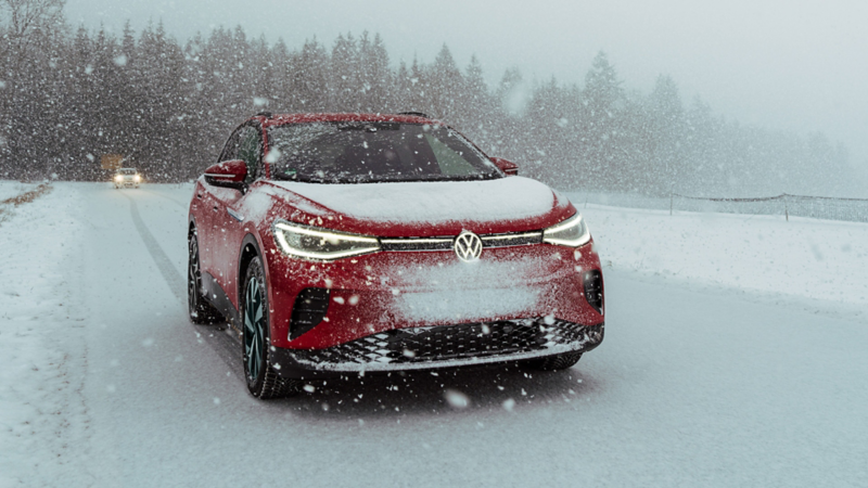 Red electric SUV driving through snowy winter conditions with headlights on, snow covering the vehicle and road, highlighting confidence and capability in harsh weather.