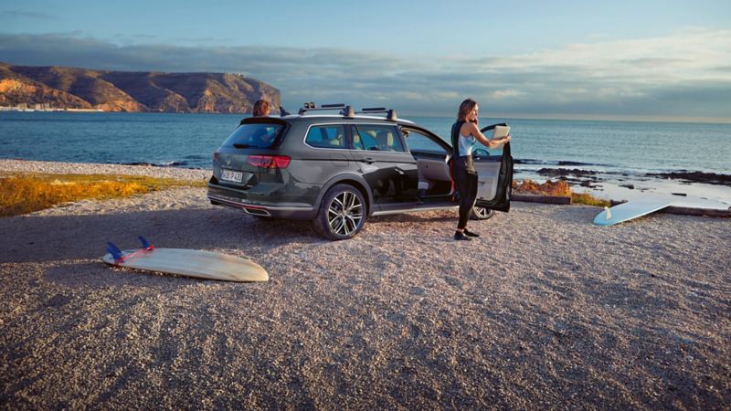 Passat Alltrack rear view on the beach, roof rack, woman standing at open passenger door, surfboard in the foreground