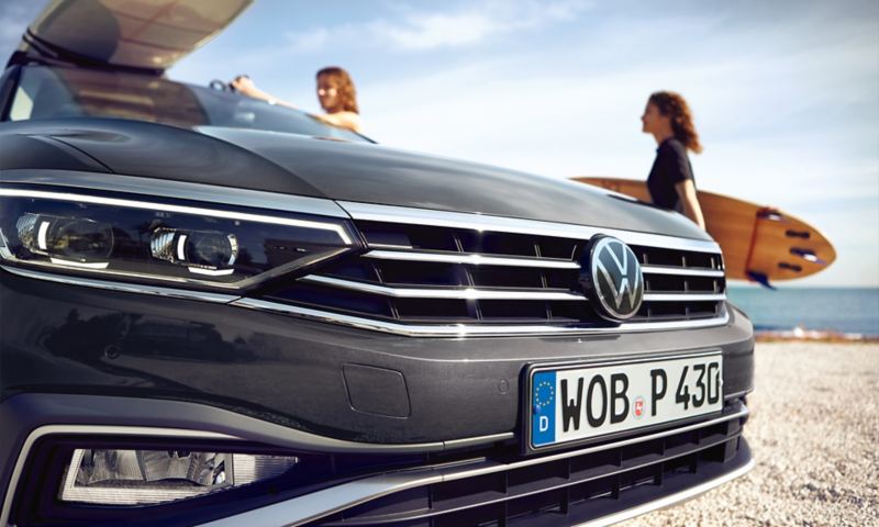 VW Passat Alltrack, Nahaufnahme Front mit Alltrack Logo im Kühlergrill, Surfbrett auf dem Dach, im Hintergrund Strand und Meer.