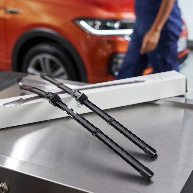  Deux balais d'essuie-glace neufs VW sont posés sur un comptoir dans un garage, avec la partie avant d'une voiture orange et une personne floue en arrière-plan.
