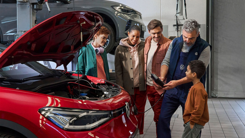 Un technicien automobile VW en uniforme bleu montre un appareil de diagnostic à un petit garçon et à un groupe de jeunes adultes dans un garage, devant une voiture rouge dont le capot est ouvert.