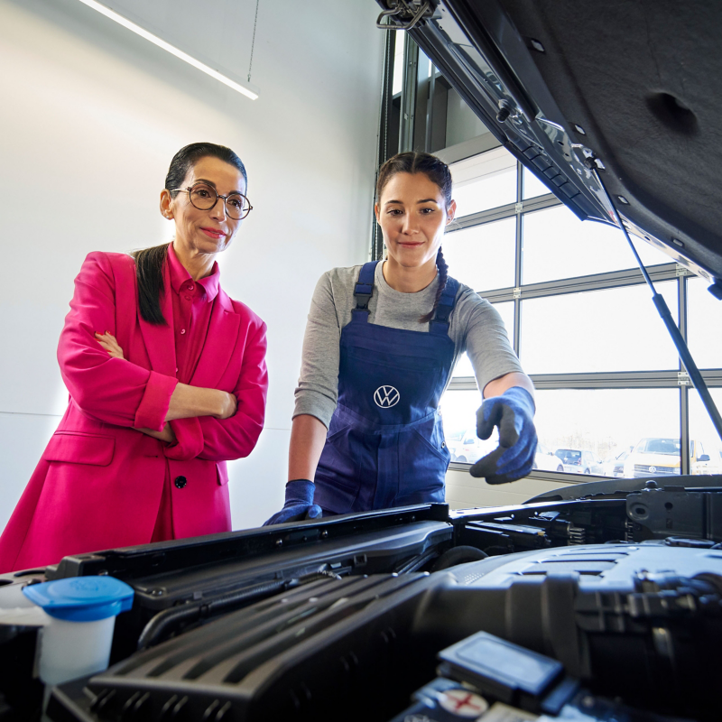  Une mécanicienne en salopette bleue explique quelque chose à une femme en blazer rose, devant un moteur de voiture ouvert dans un garage.
