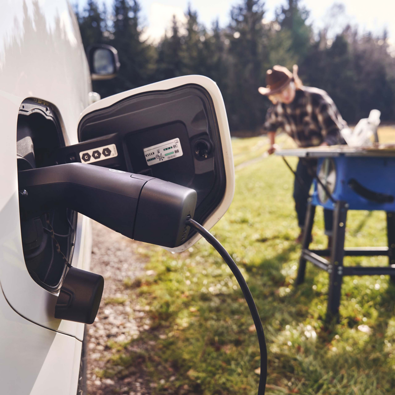 PowerSocket on VW ID. car; man wearing cowboy hat in the background, cutting wood with circular saw