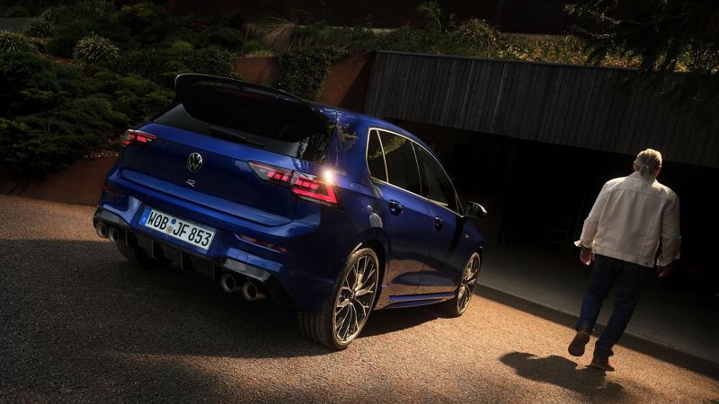 Vue de l’arrière d’une VW Golf R garée devant un garage ouvert, un homme se tient à côté