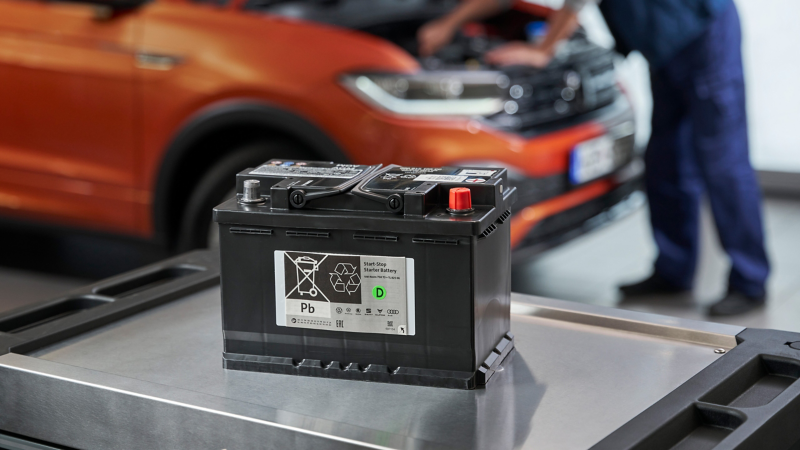  Une batterie VW de voiture noire est posée sur un établi en métal, avec une voiture orange dont le capot est ouvert et un mécanicien en arrière-plan flou.