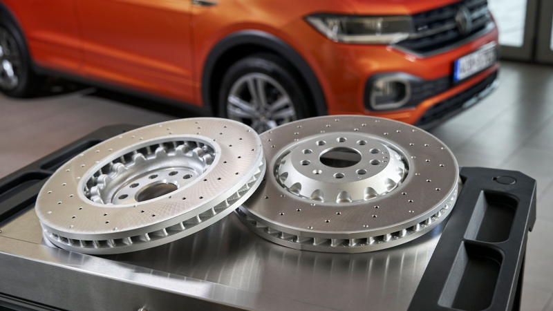  Deux disques de frein VW neufs sont posés sur un établi en métal, avec l'avant d'une voiture orange en arrière-plan flou.