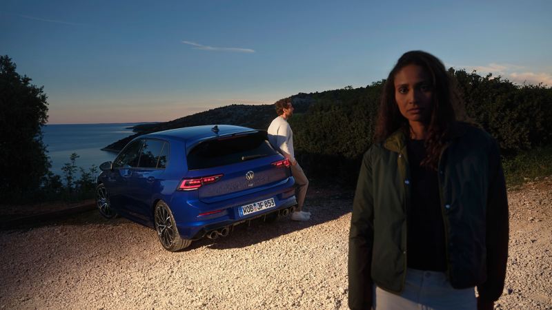 Un homme s’appuie contre une VW Golf R, la mer en arrière-plan et une femme au premier plan