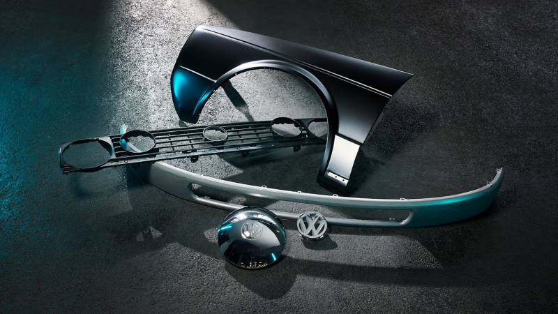 Des pièces automobiles Volkswagen classiques, dont un pare-chocs, une calandre et des logos Volkswagen, sont disposées sur une surface foncée et brillante, éclairées par une lumière bleutée.